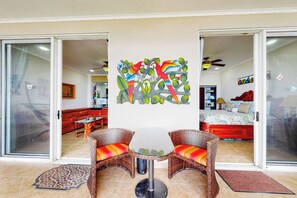 Un salón con puerta de vidrio que da a un dormitorio, una mesa de vidrio con sillas de mimbre y una pieza de arte de pared colorida.