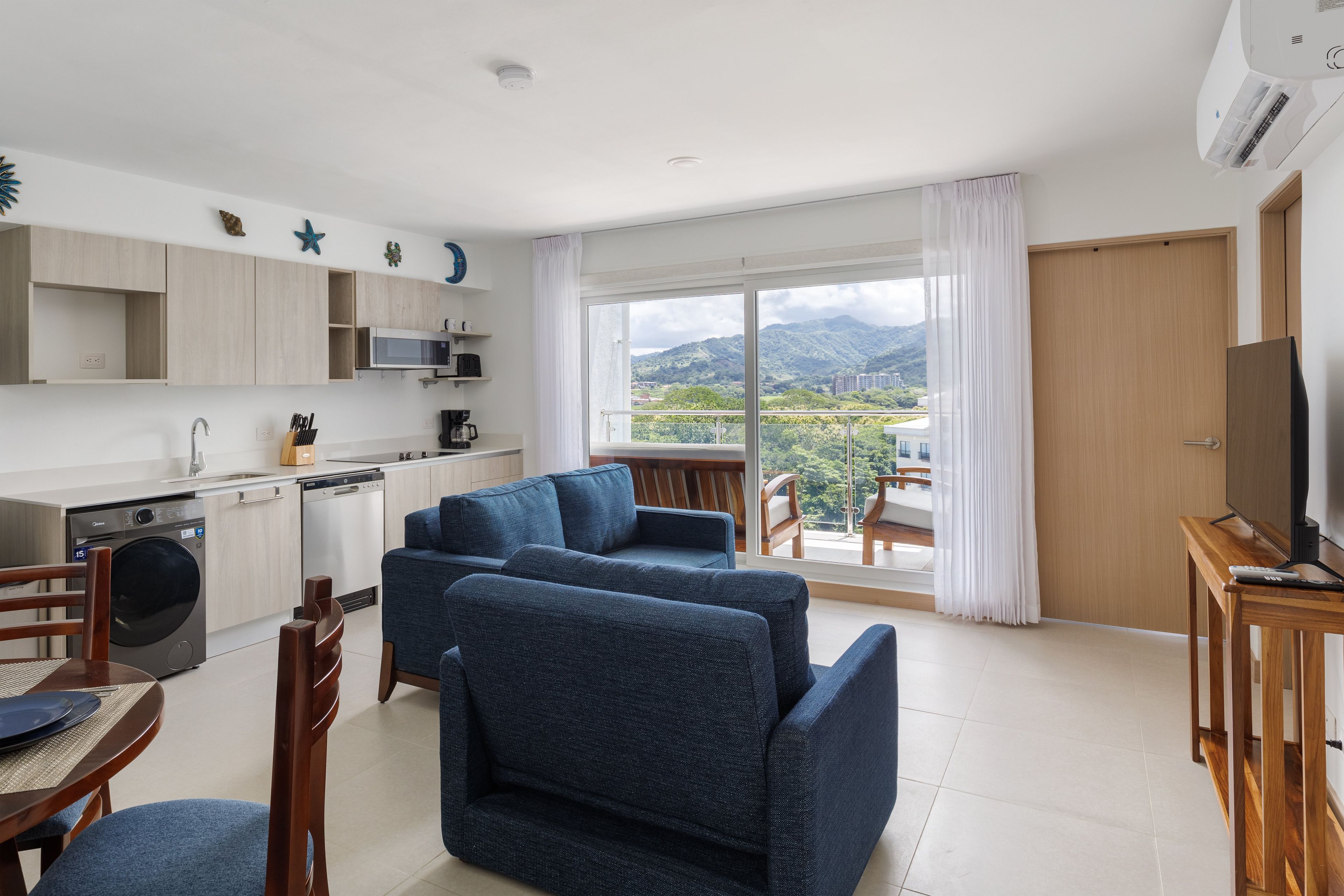 A modern living room with a sofa, dining table, and chairs, a kitchen with appliances, and a balcony with a view of mountains.