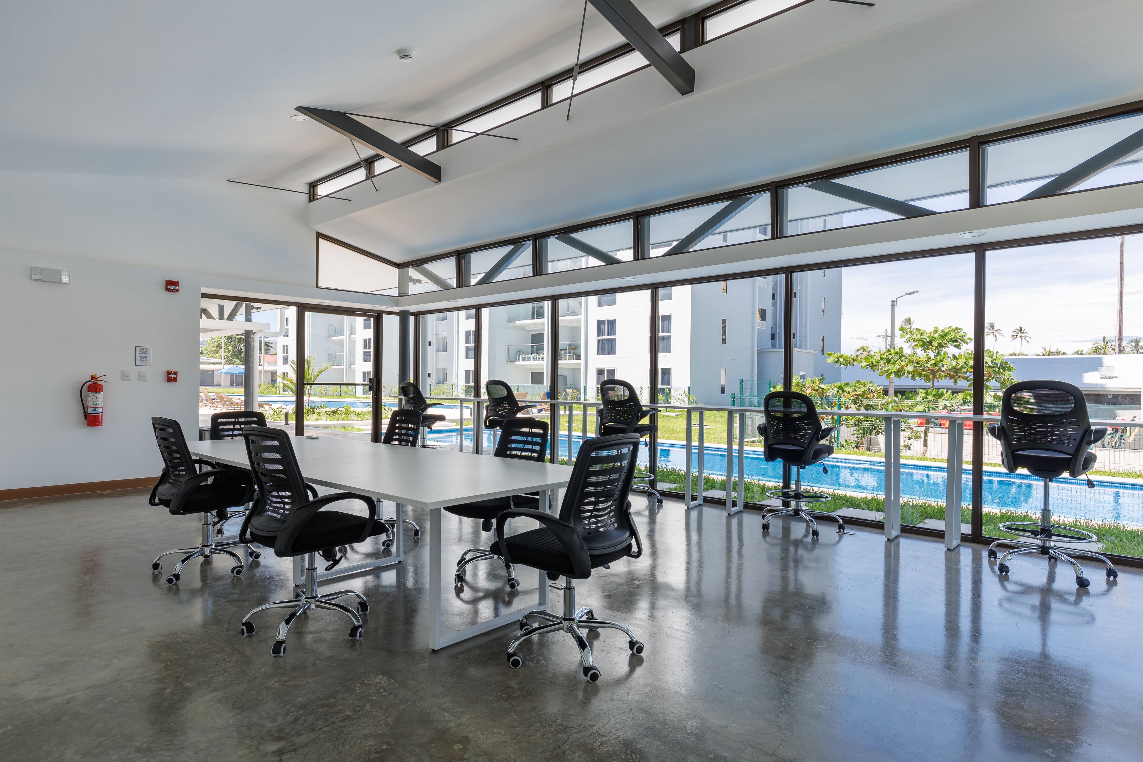 A spacious conference room with a long table and multiple chairs, large windows offering views of a swimming pool and buildings, and a fire extinguisher on the wall.