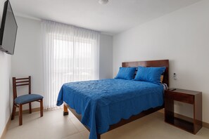 A bedroom with a bed, a chair, a bedside table, and a TV mounted on the wall. - Viva Jaco 1605 Modern Mountain View Apartment (Jacó)