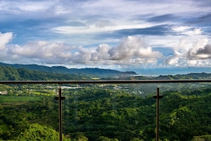 A panoramic view of a lush green landscape with mountains in the distance, viewed from a balcony with a metal railing. - Vista Serenidad 09-04C (Tarcoles)