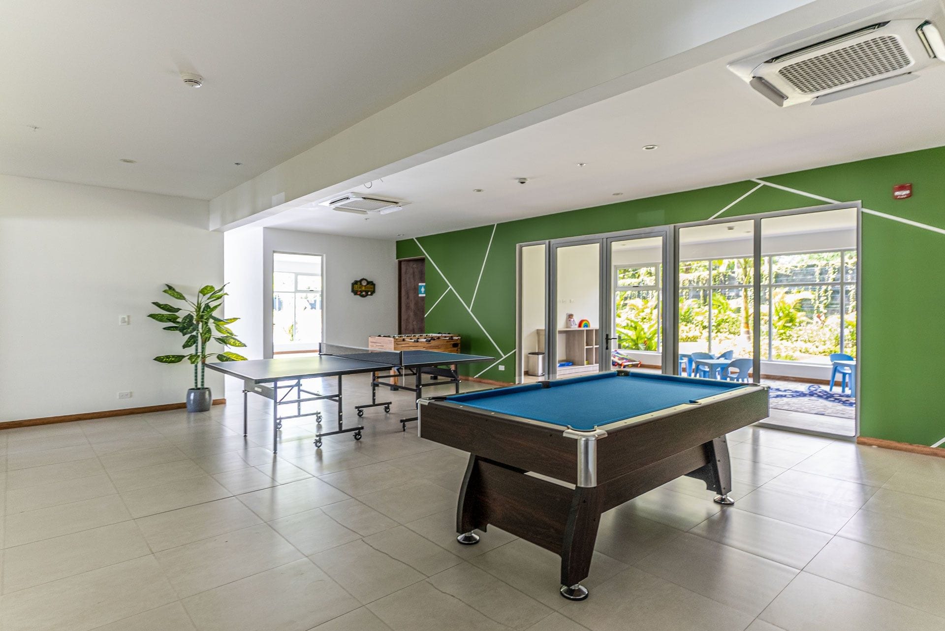 Une table de billard dans une pièce dont un mur est peint en vert, avec une plante en pot et une porte donnant sur un espace extérieur aménagé avec des chaises.