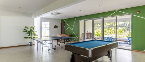 Une table de billard dans une pièce dont un mur est peint en vert, avec une plante en pot et une porte donnant sur un espace extérieur aménagé avec des chaises.