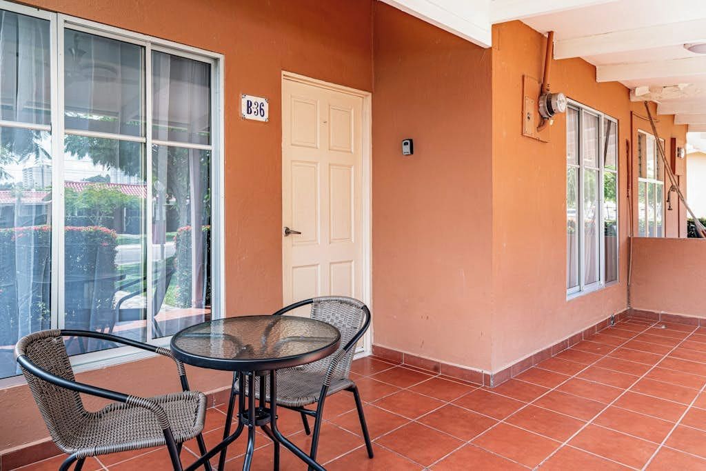 Une terrasse couverte avec une table et des chaises, une porte avec une pancarte et une fenêtre donnant sur la verdure.