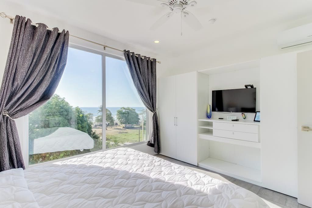 Ein Schlafzimmer mit einem großen Bett, einem Fernseher auf einer weißen Unterhaltungsanlage und einer Glastür, die auf einen Balkon mit Blick gelangt.