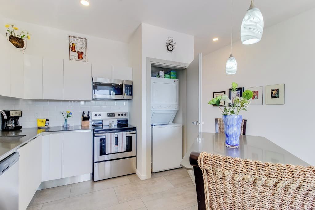 A modern kitchen with white cabinets, a built-in oven, microwave, and refrigerator. There is a dining area with a woven chair and a vase of flowers on the table.