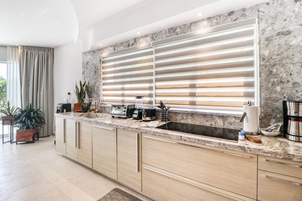 A modern kitchen with a marble countertop, built-in oven, and a large window with blinds.