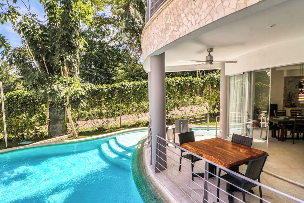 Una piscina con tavolo e sedie in legno, immersa nel verde e affiancata da un edificio moderno con ampie vetrate.