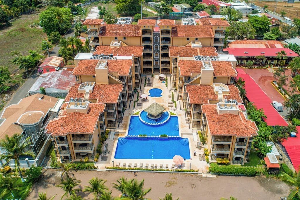 Un hotel a più piani con piscina centrale, circondato da balconi e un’area parcheggio con tetto rosso.