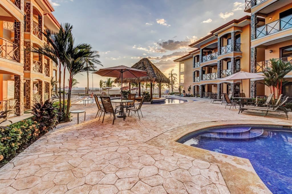 A resort pool area with hexagonal tiles, lounge chairs, umbrellas, and a thatched-roof structure.
