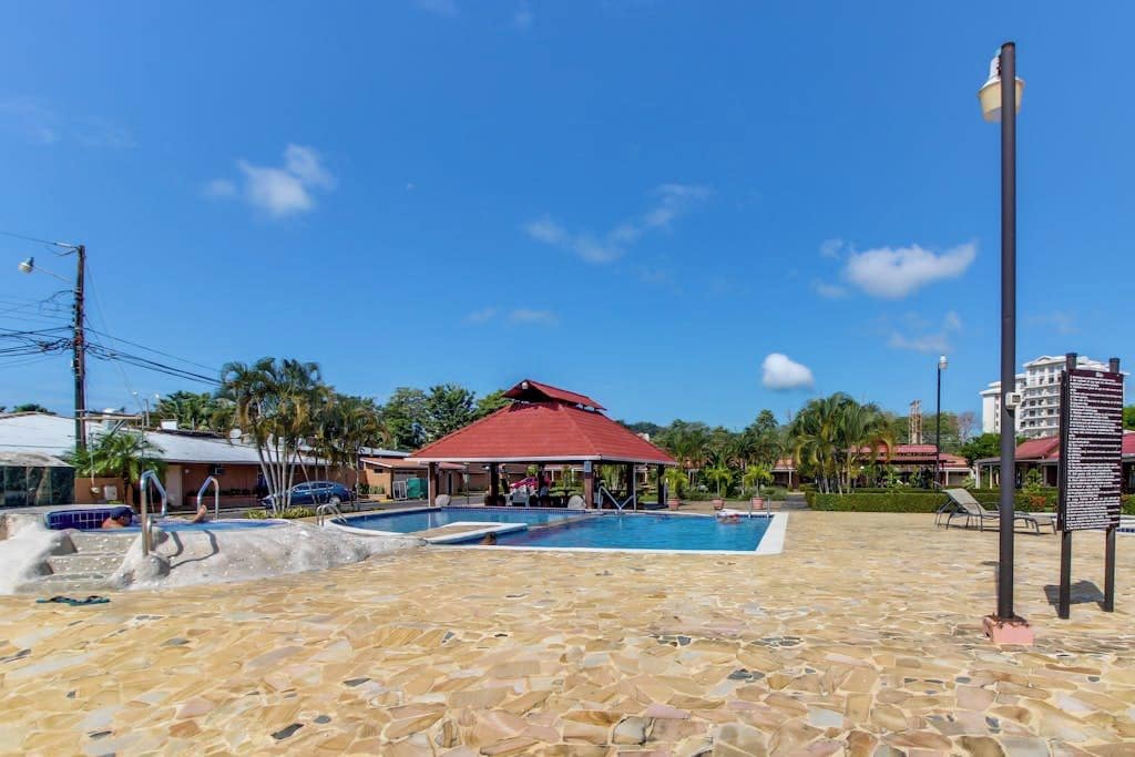 A swimming pool area with a red-roofed pavilion, lounge chairs, and a tiled deck.