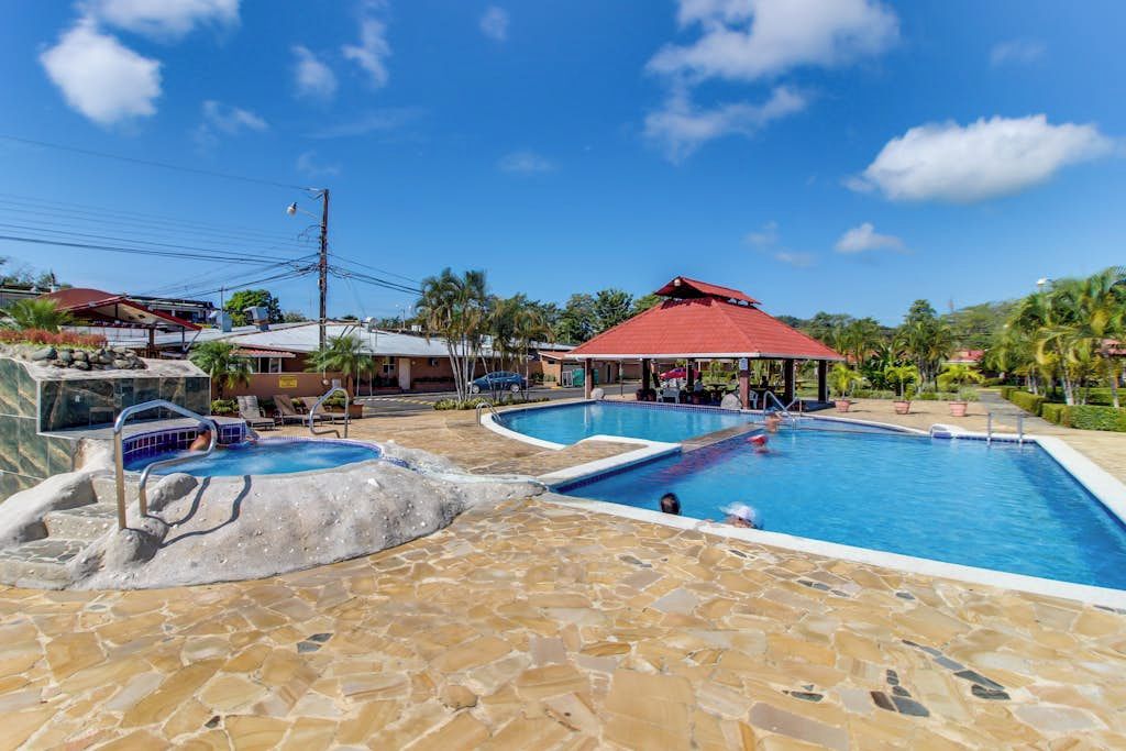 A swimming pool area with a stone floor, a red-roofed pavilion, and a clear blue sky.