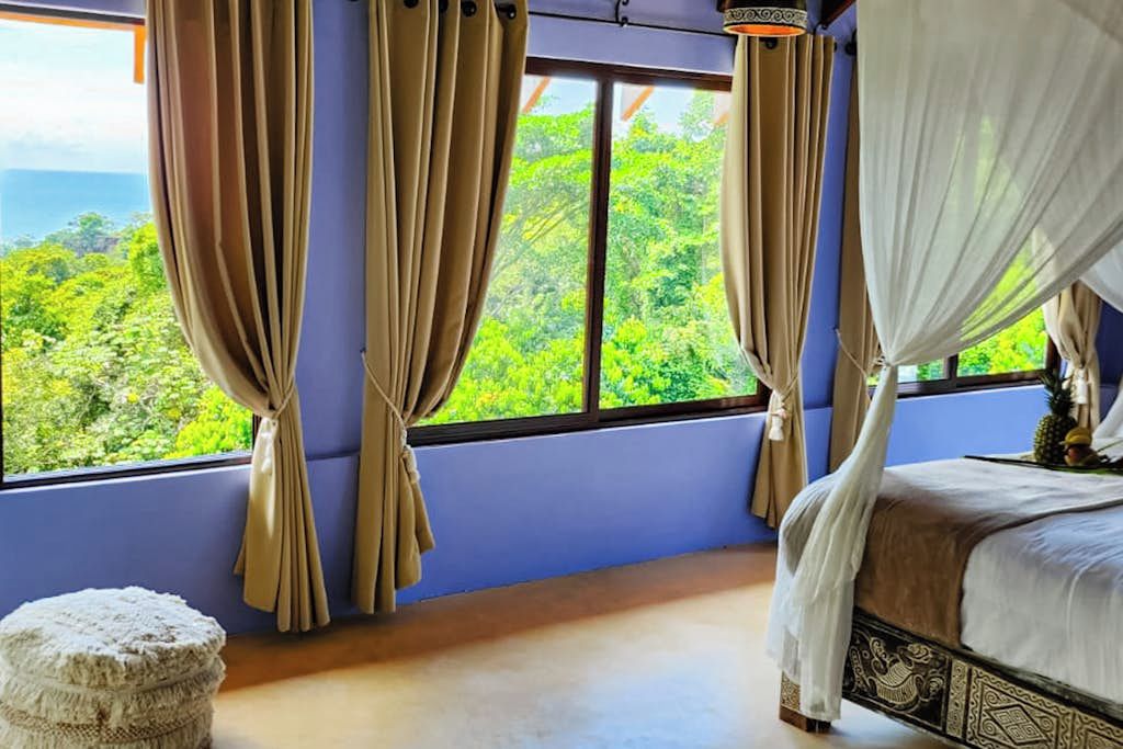 A bedroom with a large window, curtains, a bed, and a small round ottoman.