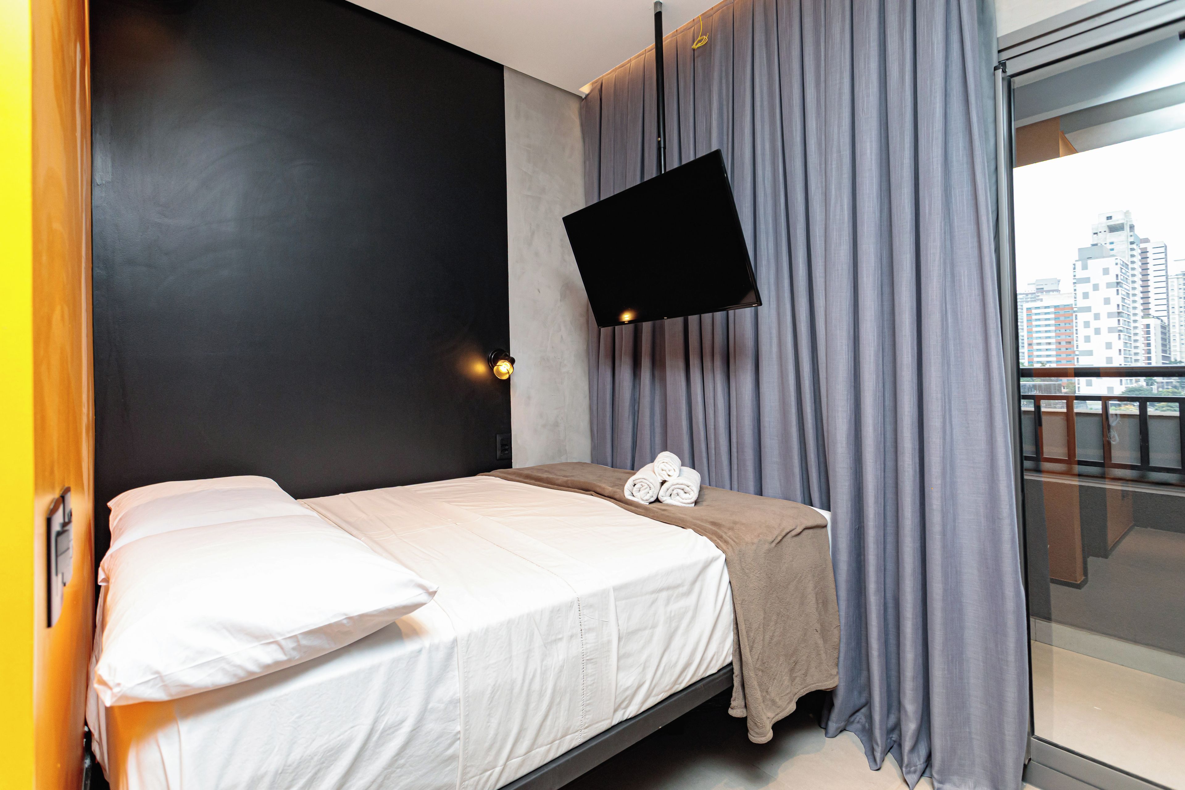 A hotel room with a bed, a flat-screen TV mounted on the wall, a window with curtains, and a view of the cityscape.