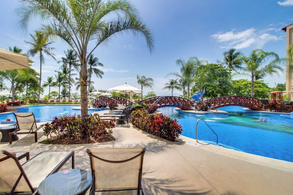 A pool area with lounge chairs, a bridge, and palm trees.