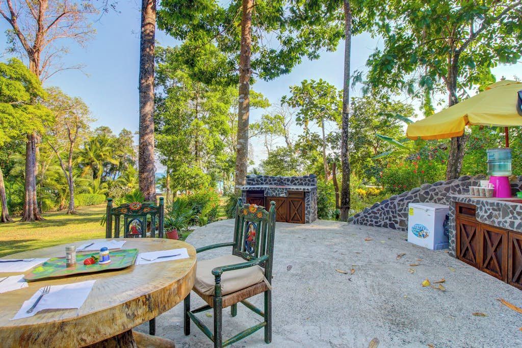 An outdoor dining area with a table and chairs, surrounded by trees and a stone structure.