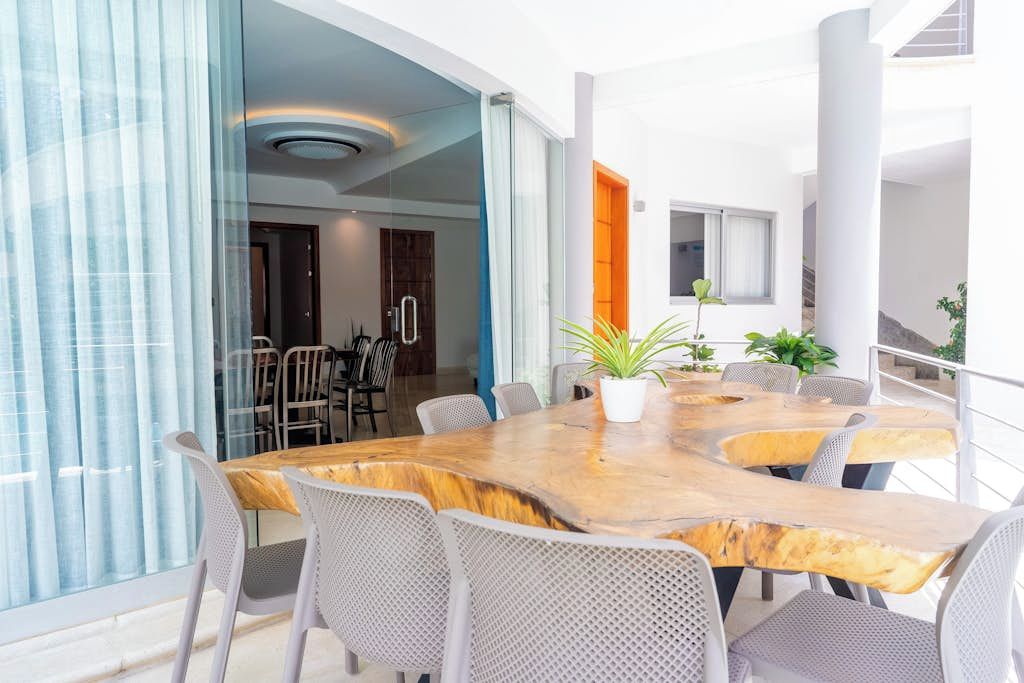 A modern dining area with a large wooden table and white chairs, a potted plant, and a view of the interior space.