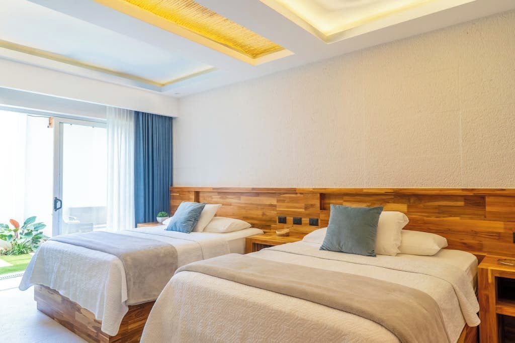 A hotel room with two beds, a wooden headboard, a large window with curtains, and a ceiling with recessed lighting.