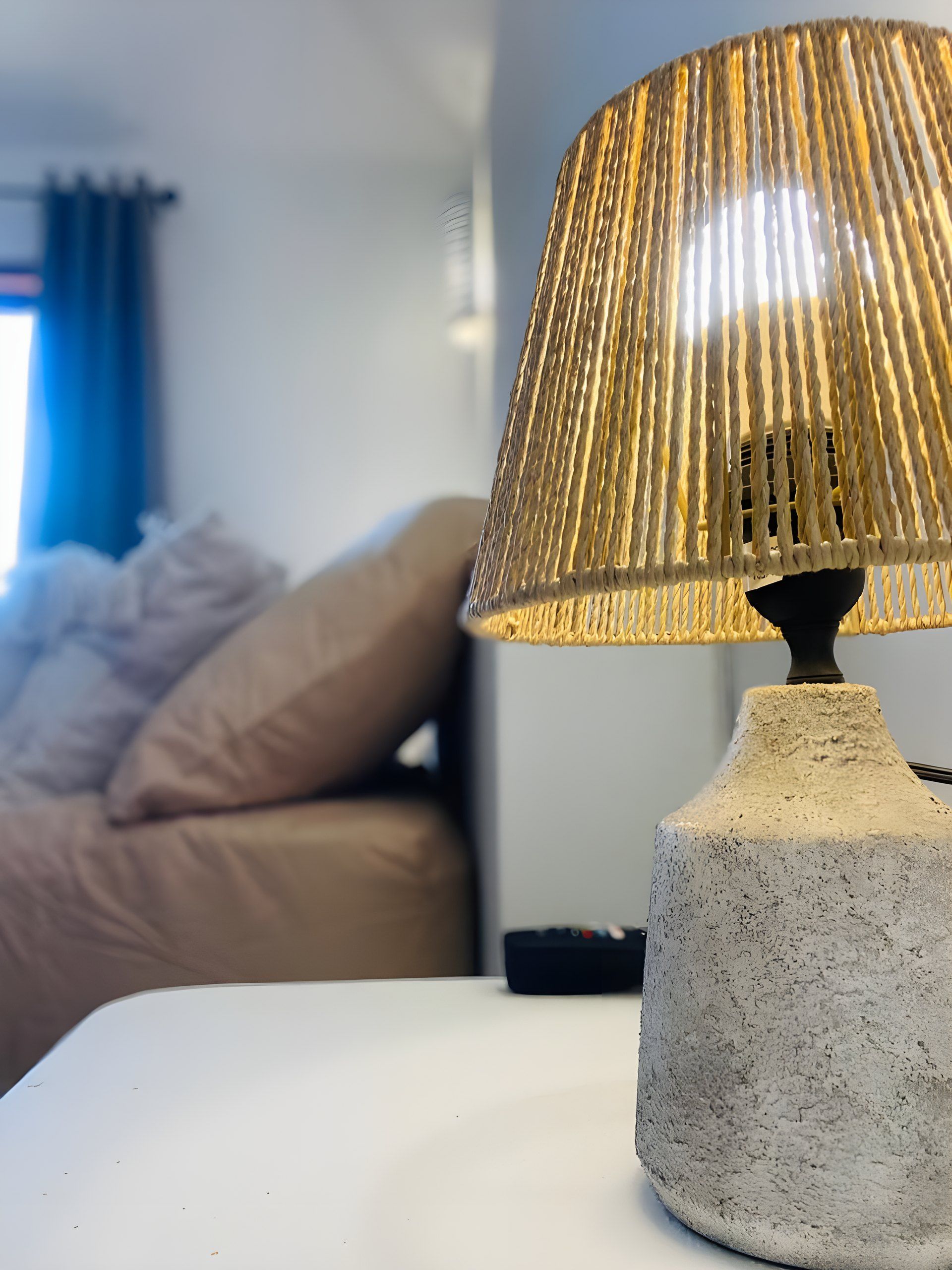 Une lampe avec un abat-jour texturé posée sur une table blanche, un lit avec une literie beige et un rideau bleu en arrière-plan.