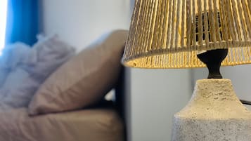Une lampe avec un abat-jour texturé posée sur une table blanche, un lit avec une literie beige et un rideau bleu en arrière-plan.