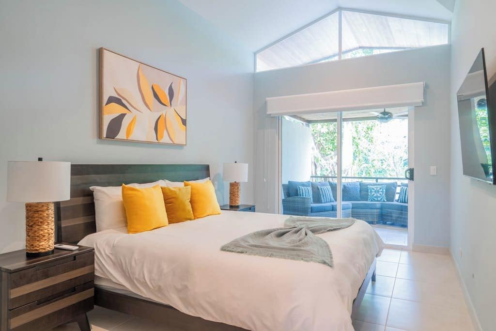A bedroom with a bed, bedside lamps, a painting, and a view of an outdoor seating area.