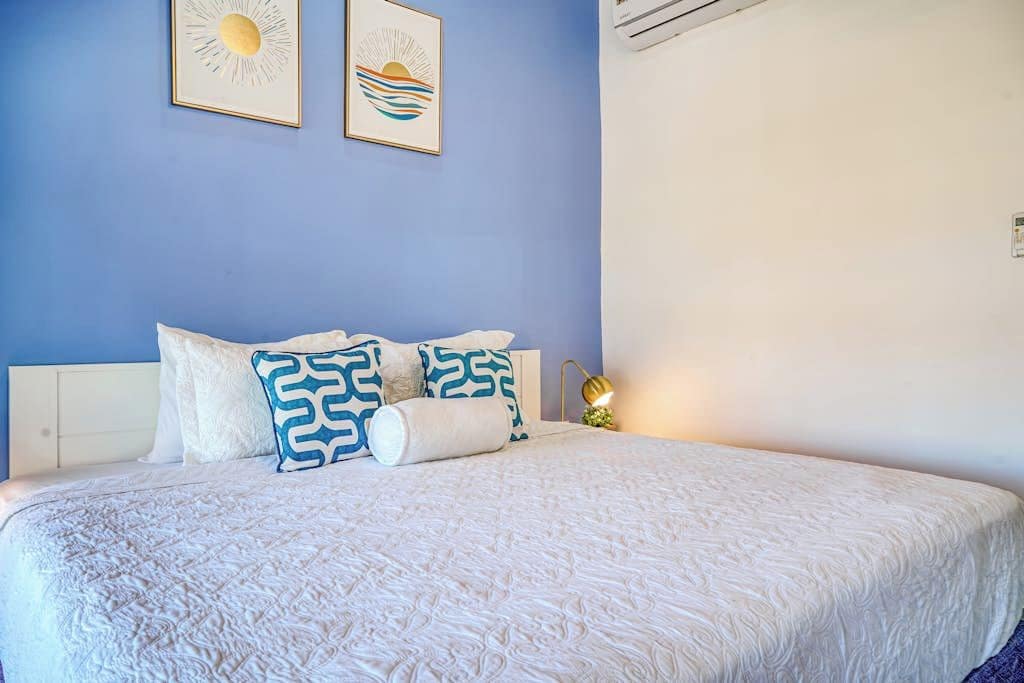 A bed with a white quilt and patterned pillows, a bedside lamp, and two framed artworks on the wall.