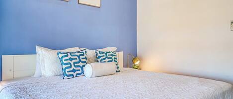 Un lit avec une couette blanche et des oreillers à motifs, une lampe de chevet et deux œuvres d’art encadrées accrochées au mur.