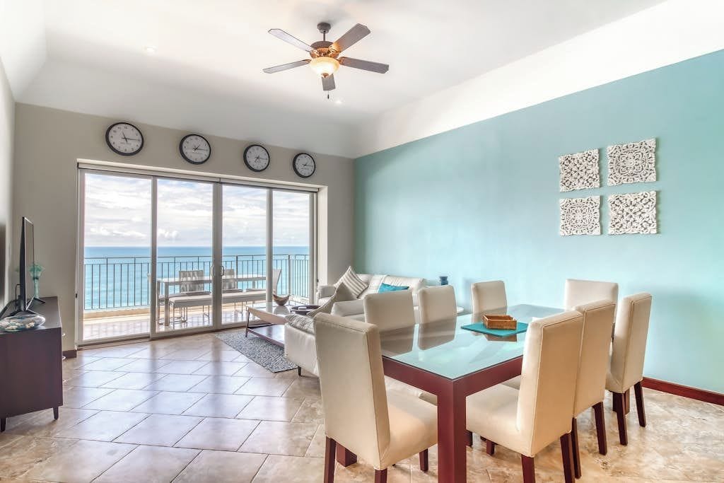 A spacious living area with a dining table, chairs, a sofa, and a balcony with a view of the ocean.