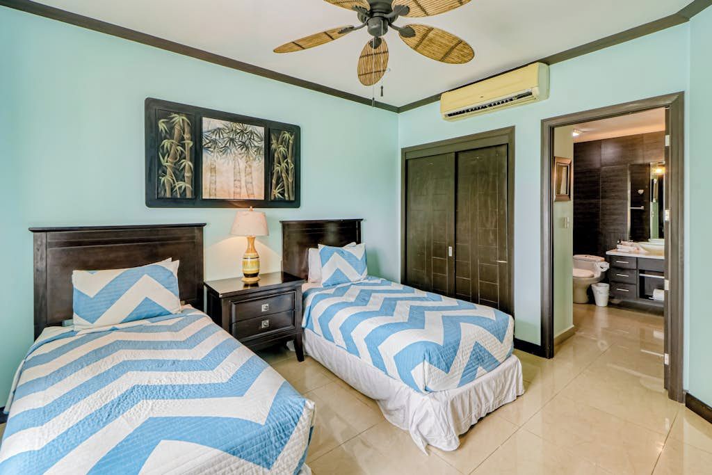 A hotel room with two beds, a ceiling fan, a nightstand with a lamp, a door leading to a bathroom, and artwork on the wall.