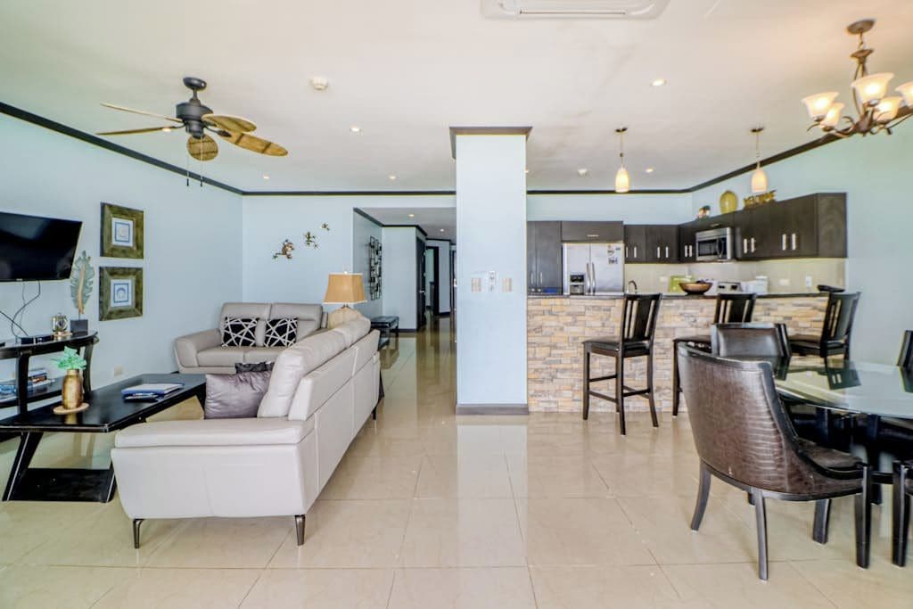 A modern living room with a sofa, a dining table with chairs, a kitchen area with cabinets, and a ceiling fan.