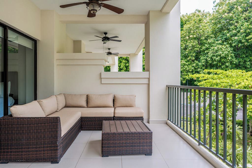 Ein Balkon mit Rattan-Sofa, Hocker und Deckenventilatoren, mit Blick auf viel Grün.