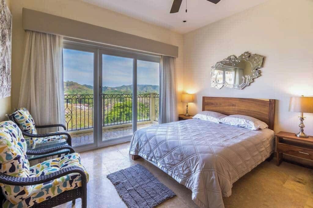 Habitación de hotel con cama, una silla, mesita de noche, lámpara, balcón con vistas y un espejo en la pared.