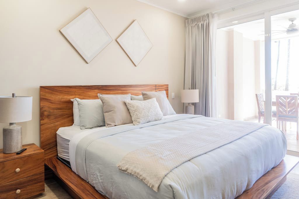 A bedroom with a wooden headboard, a large bed with multiple pillows, a bedside table with a lamp, and a view through a glass door to an outdoor area.