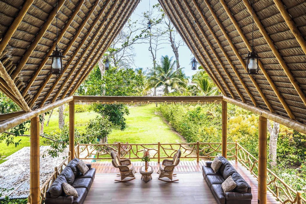A covered outdoor seating area with a thatched roof, furnished with sofas and chairs, overlooking a well-maintained garden.