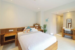Une chambre d’hôtel comprenant un lit, des tables de chevet, une petite table de nuit, une salle de bain avec un lavabo et un miroir, et une petite plante en pot.