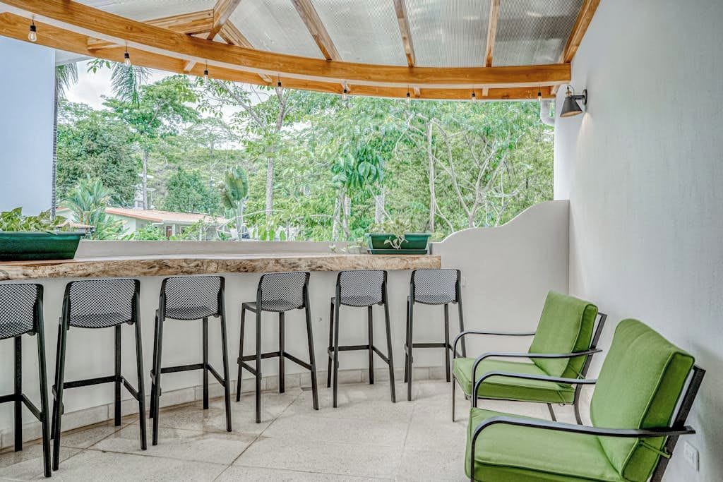 A covered outdoor seating area with a bar, chairs, and a view of greenery.