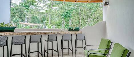 A covered outdoor seating area with a bar, chairs, and a view of greenery.