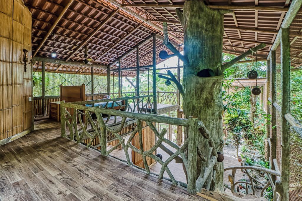 An indoor wooden structure with a thatched roof, a wooden bench, and a tree trunk with hanging objects.