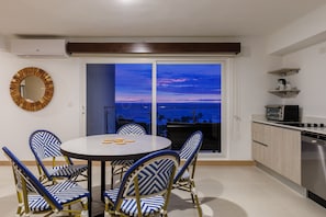 Ein Essbereich mit rundem Tisch und blau-goldenen Stühlen, eine Küche mit Geschirrspüler und Blick auf den Ozean im Abendrot.