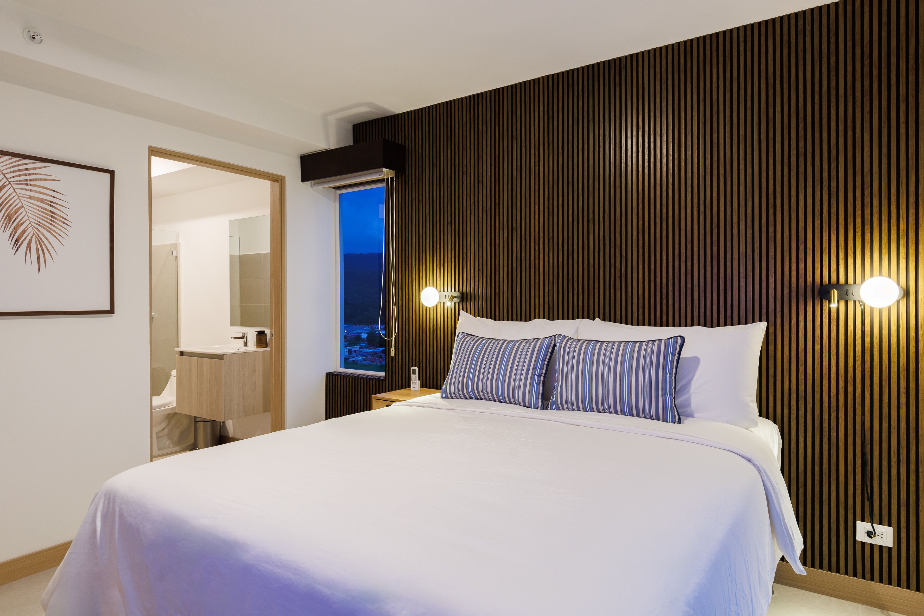 A modern hotel room with a large bed, wooden panel wall, bedside lamps, and a view of the city at dusk.