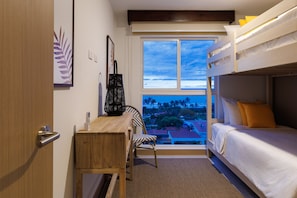 Ein Zimmer mit Etagenbett, Schreibtisch und Stuhl. Ein Fenster bietet Blick auf Palmen und Gebäude.