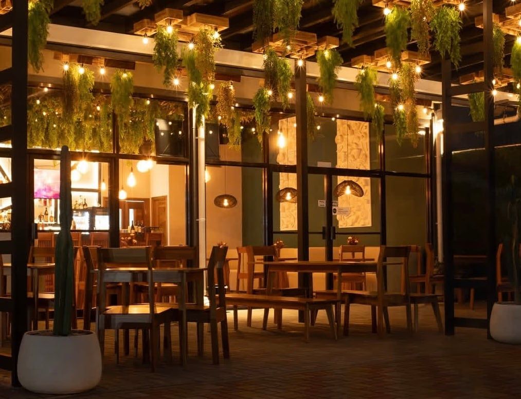 An indoor dining area with wooden tables and chairs, hanging plants, and warm lighting.