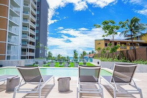 A poolside area with lounge chairs, a clear sky, and a view of a building with balconies. - Pacific Point 705 Beachfront Condo in Jaco (Jaco)