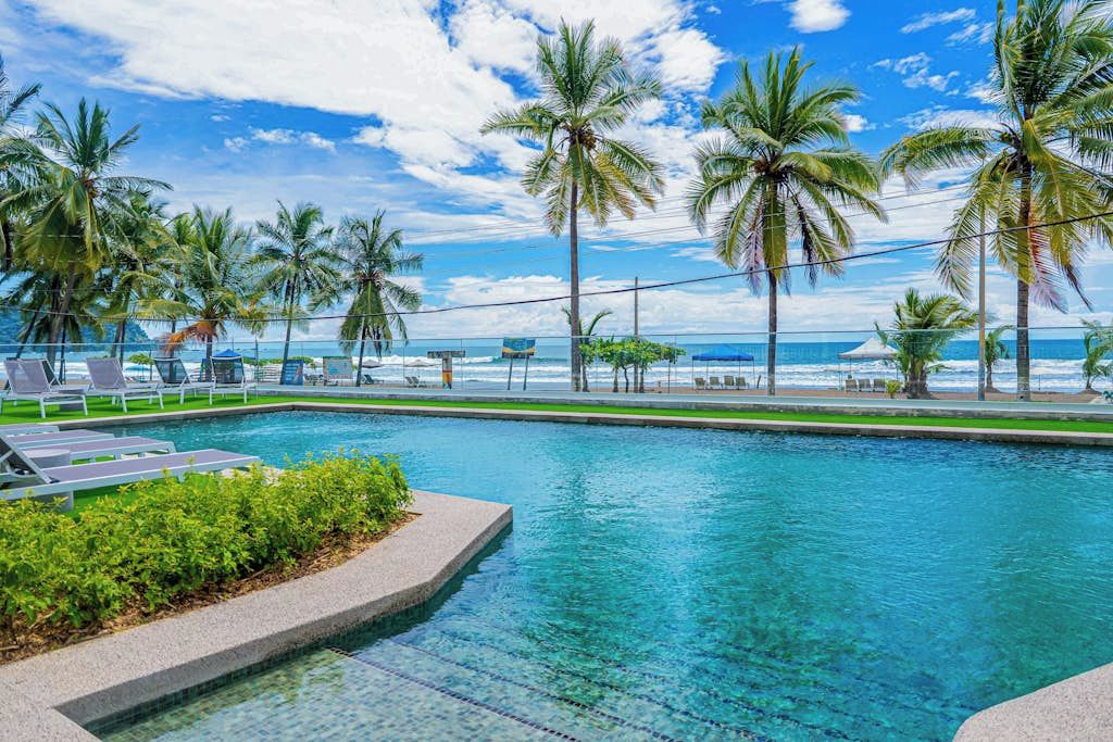 A pool with clear blue water, surrounded by palm trees and lounge chairs, with a view of the ocean.