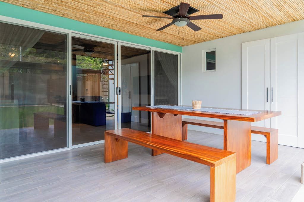 Une table en bois avec des bancs, dans une pièce dotée d’un ventilateur de plafond, de portes-fenêtres coulissantes et offrant une vue sur l’extérieur.