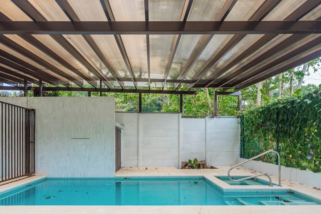 Una piscina con zona cubierta, rodeada de un muro de hormigón y vegetación.