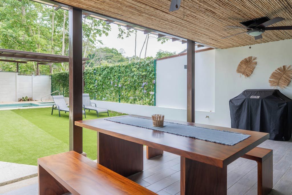 Ein moderner Essbereich im Freien mit Holztisch und -bänken, einer Pergola und Blick auf einen Swimmingpool und Grünflächen.
