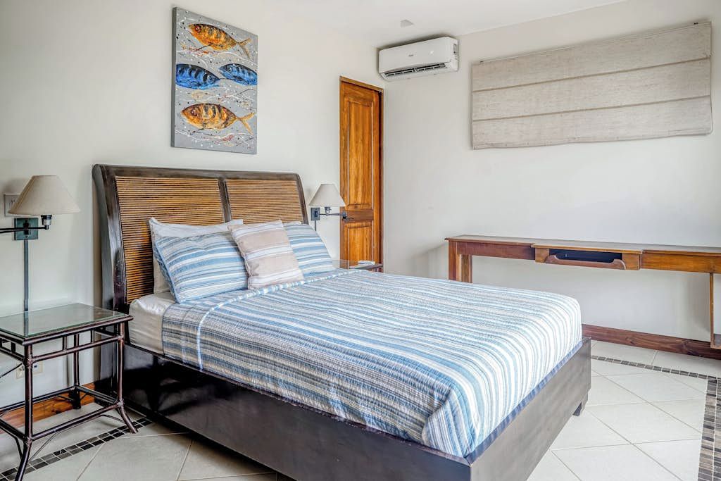 Une chambre à coucher comprenant un lit, des tables de chevet, une commode, une tête de lit en bois, un climatiseur mural, une porte en bois et une œuvre d’art encadrée.
