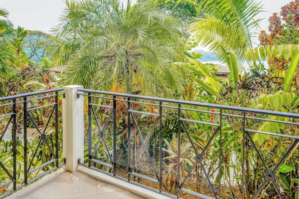 Un balcon doté d’une balustrade en métal, donnant sur un jardin luxuriant où poussent différentes plantes tropicales, et offrant une vue sur la mer.