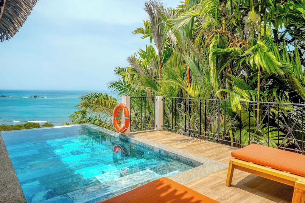 Ein Infinity-Pool mit freiem Blick auf das Meer, umgeben von üppiger tropischer Vegetation und einer Holzterrasse mit einer Liege.